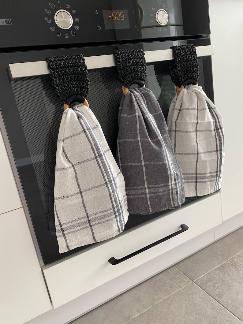 May include: Three kitchen towels hanging on a stainless steel bar. The towels have a plaid pattern in shades of gray and white. Each towel is held by a black crocheted loop and a wooden ring. The towels are hanging on a black oven.