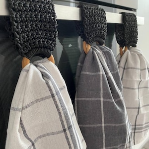 May include: Three kitchen towel holders with black macrame detailing, each holding a plaid kitchen towel. The towels are in shades of white and grey. The holders have wooden rings and hang from a stainless steel bar.