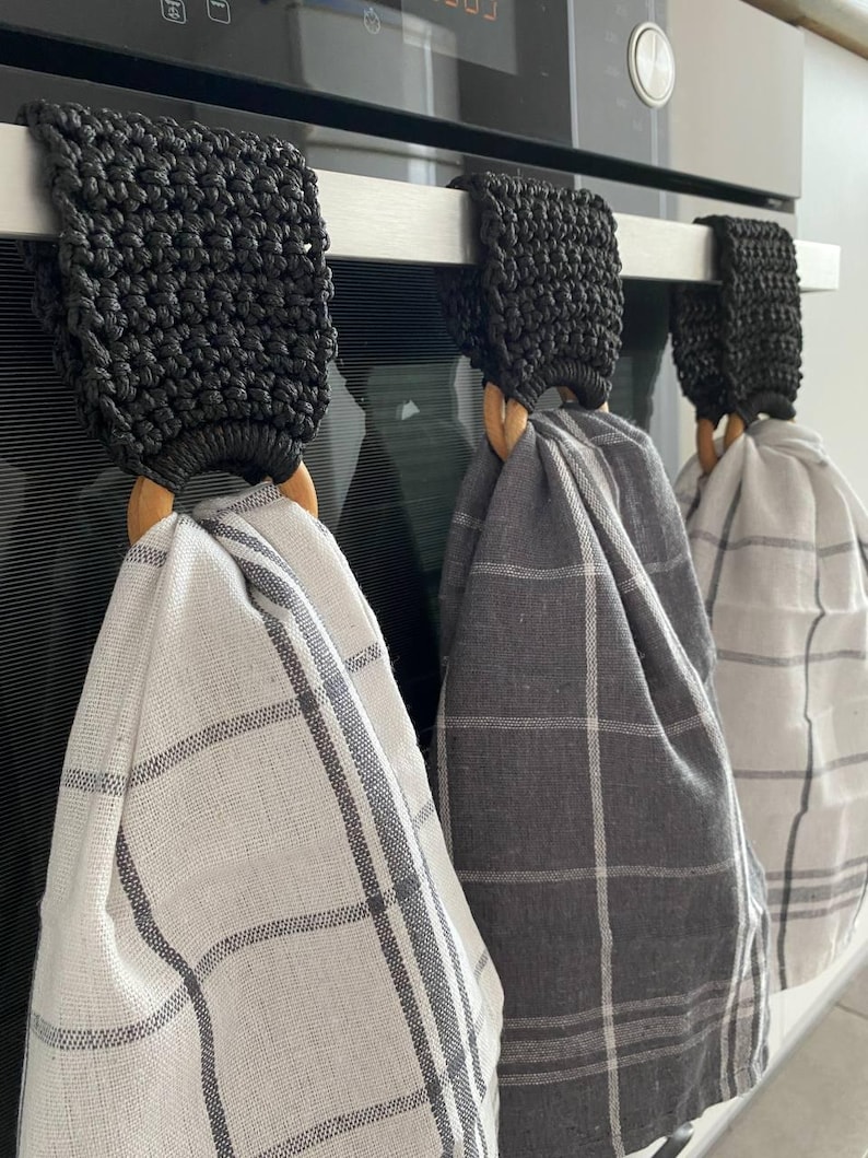 May include: Three kitchen towel holders with macrame detailing in black, each holding a plaid kitchen towel. The towels are in shades of white and gray. The holders have wooden rings and hang from a stainless steel bar.