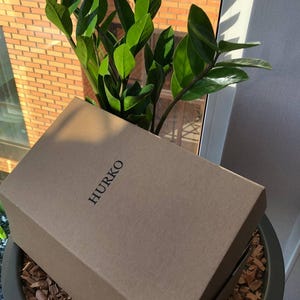 K&ouml;nnte beinhalten: Ein brauner Pappkarton mit dem Wort "HURKO" auf der Oberseite. Die Box liegt auf einer Schicht aus Holzsp&auml;nen in einem gr&uuml;nen Topf. Im Hintergrund befindet sich eine gr&uuml;ne Pflanze mit gro&szlig;en Bl&auml;ttern.