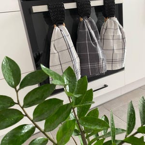 May include: Three kitchen towels with black crochet loops hanging on an oven handle. The towels have a plaid pattern in shades of gray and white. A green plant with glossy leaves is in the foreground.