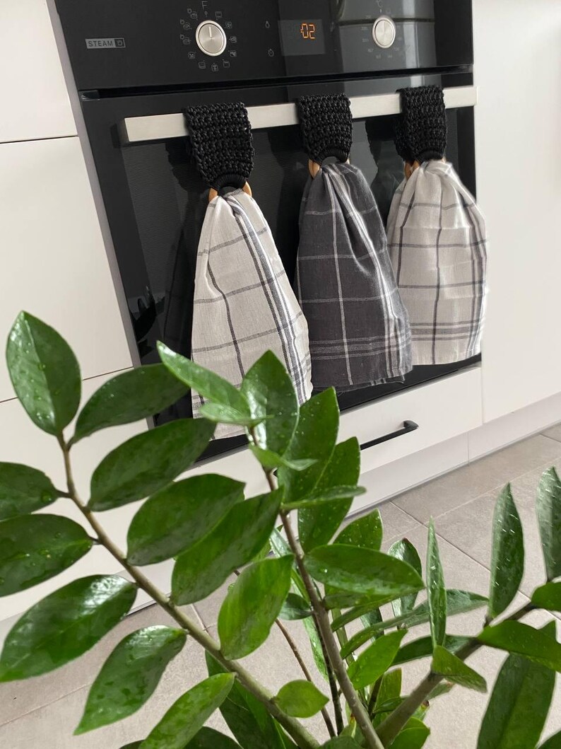 May include: Three kitchen towels with black crochet loops hanging on an oven handle. The towels have a plaid pattern in shades of gray and white. A green plant with glossy leaves is in the foreground.