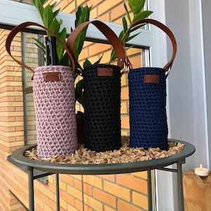May include: Three crochet wine bottle holders in pink, black, and navy blue, each with a brown leather handle and a small leather tag. The holders are displayed on a round metal table with wood chips.