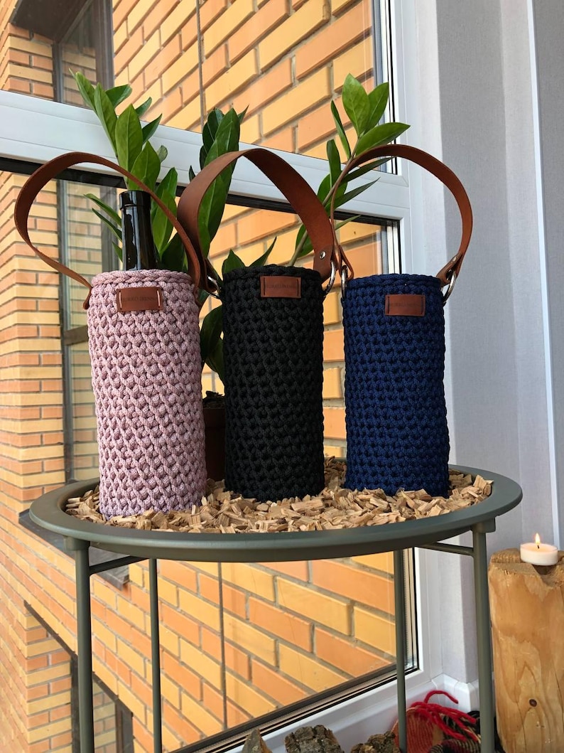 K&ouml;nnte beinhalten: Drei H&auml;kel-Weinflaschenhalter in Rosa, Schwarz und Marineblau, jeweils mit einem braunen Ledergriff und einem kleinen Lederetikett. Die Halter werden auf einem runden Metalltisch mit Holzsp&auml;nen ausgestellt.