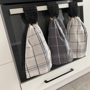 May include: Three kitchen towels hanging on an oven. Each towel is made of a plaid fabric in shades of gray and white. The towels are held by wooden rings and black crocheted loops.
