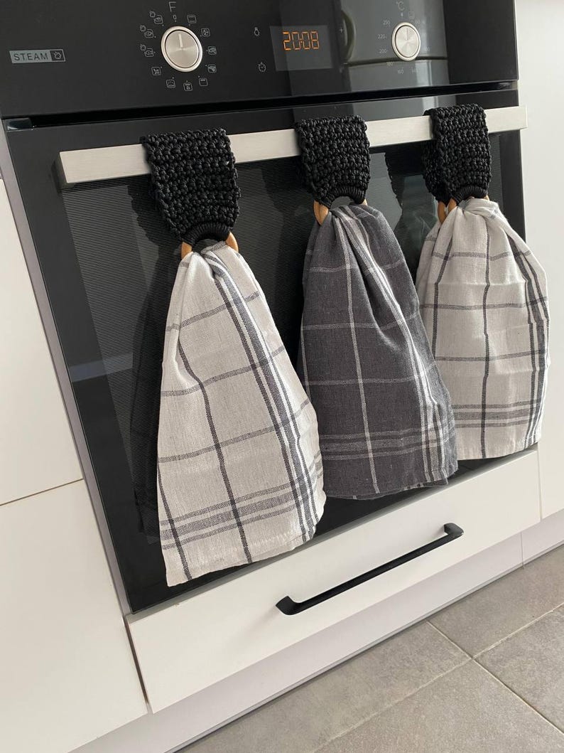 May include: Three kitchen towels hanging on an oven. Each towel is made of a plaid fabric in shades of gray and white. The towels are held by wooden rings and black crocheted loops.