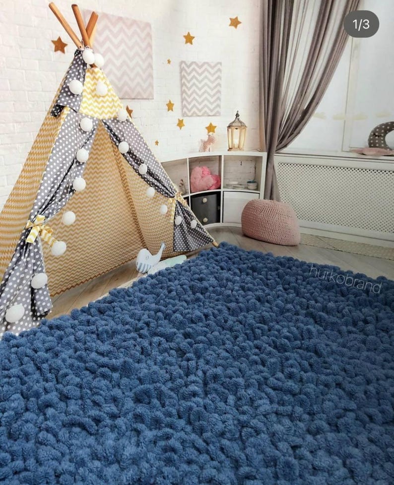 Peut inclure: Une chambre d'enfant confortable avec un tapis bleu textur&eacute;, un tipi avec des motifs jaunes et gris, et des &eacute;toiles d&eacute;coratives sur le mur en briques blanches. La pi&egrave;ce comprend une petite &eacute;tag&egrave;re avec des jouets et un pouf rose.