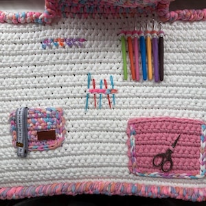 Puede incluir: Un organizador de manualidades de ganchillo blanco con ribetes rosas, azules y morados. Cuenta con bolsillos para tijeras, una cinta métrica y otros suministros de manualidades. También se ven ganchos y agujas de ganchillo.