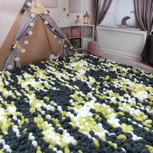 Peut inclure: Un tapis fait &agrave; la main avec une texture de pompons dans une chambre d'enfant. Le tapis pr&eacute;sente un motif de pompons gris fonc&eacute;, blancs et vert clair. Une tente tipi avec des pois gris et un motif chevron beige est visible.
