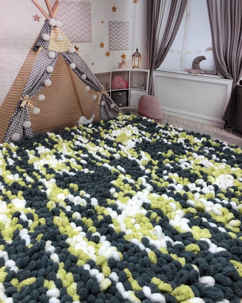 Peut inclure: Un tapis fait &agrave; la main avec une texture de pompons dans une chambre d'enfant. Le tapis pr&eacute;sente un motif de pompons gris fonc&eacute;, blancs et vert clair. Une tente tipi avec des pois gris et un motif chevron beige est visible.