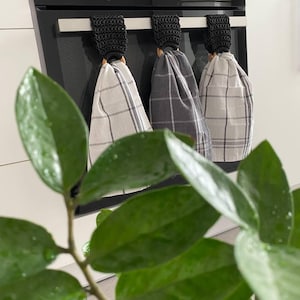 May include: Three kitchen towels with plaid patterns hang from a black oven. The towels are in shades of gray and white. Each towel is held by a black crocheted loop with a wooden ring. A green plant is in the foreground.