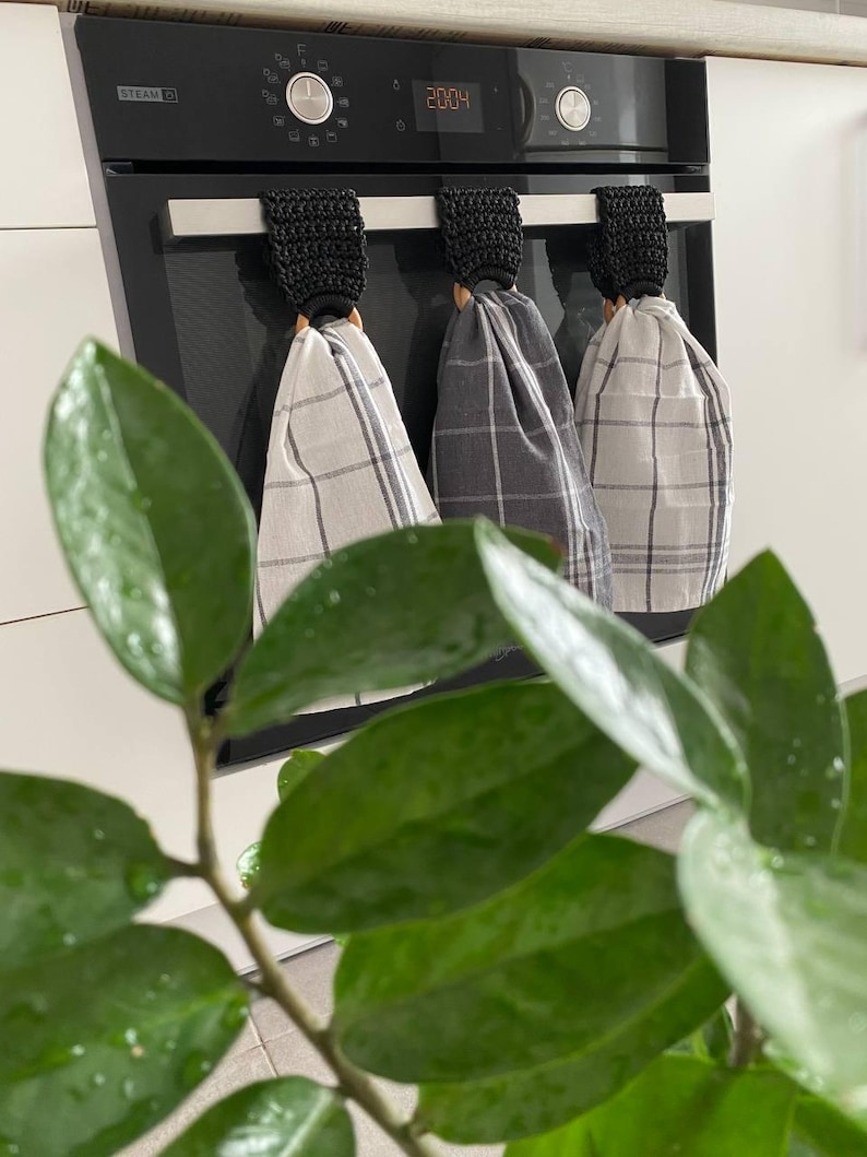May include: Three kitchen towels with plaid patterns hang from a black oven. The towels are in shades of gray and white. Each towel is held by a black crocheted loop with a wooden ring. A green plant is in the foreground.