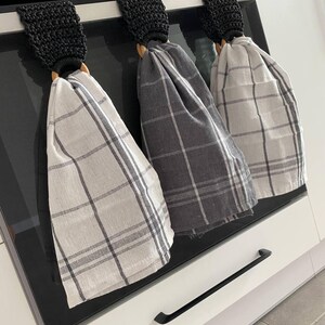 May include: Three kitchen towels hanging on an oven. The towels are gray and white plaid. Each towel is held by a wooden ring and a black crocheted loop. The oven is black with a digital clock.