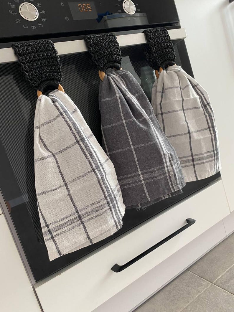 May include: Three kitchen towels hanging on an oven. The towels are gray and white plaid. Each towel is held by a wooden ring and a black crocheted loop. The oven is black with a digital clock.