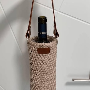 K&ouml;nnte beinhalten: Ein beigefarbener, geh&auml;kelter Weinflaschenhalter mit braunem Ledergriff und Akzenten. Der Halter hat eine zylindrische Form und ist f&uuml;r eine einzelne Weinflasche konzipiert. Die Flasche im Inneren tr&auml;gt die Aufschrift "Sherel".