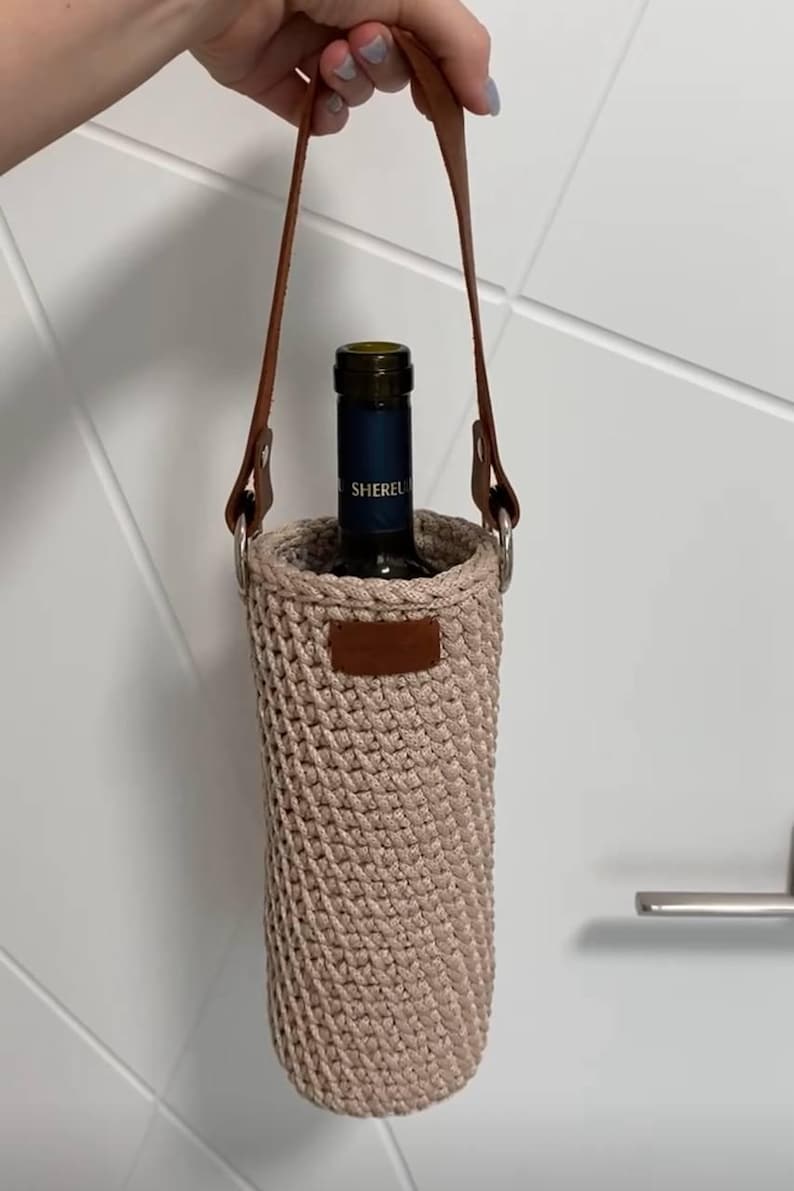 K&ouml;nnte beinhalten: Ein beigefarbener, geh&auml;kelter Weinflaschenhalter mit braunem Ledergriff und Akzenten. Der Halter hat eine zylindrische Form und ist f&uuml;r eine einzelne Weinflasche konzipiert. Die Flasche im Inneren tr&auml;gt die Aufschrift "Sherel".