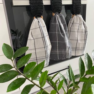 May include: Three kitchen towels with black crochet tops and wooden rings hanging on a stainless steel bar. The towels are in a plaid pattern, with colors of gray and white. A green plant with large leaves is in the foreground.