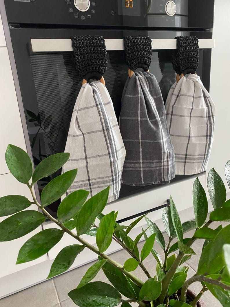 May include: Three kitchen towels with black crochet tops and wooden rings hanging on a stainless steel bar. The towels are in a plaid pattern, with colors of gray and white. A green plant with large leaves is in the foreground.