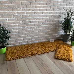 Peut inclure: Ensemble de tapis en tricot &eacute;pais jaune moutarde. L'ensemble comprend un long tapis rectangulaire et un plus petit tapis carr&eacute;. Les tapis sont pos&eacute;s sur un plancher en bois clair, avec des plantes en pot et un mur de briques en arri&egrave;re-plan.