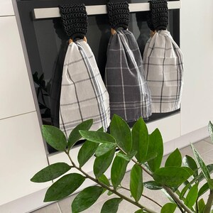 May include: Three kitchen towels with plaid patterns hang from a black oven handle. The towels are gray and white, and the holders are black crochet with wooden rings. A green plant with glossy leaves is in the foreground.