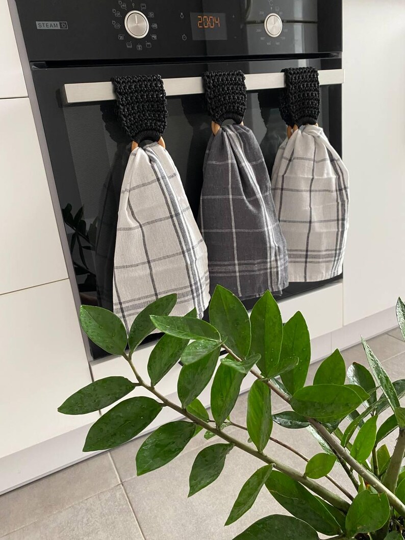 May include: Three kitchen towels with plaid patterns hang from a black oven handle. The towels are gray and white, and the holders are black crochet with wooden rings. A green plant with glossy leaves is in the foreground.