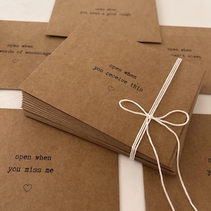 May include: A stack of brown kraft paper envelopes with white string tied around them. Each envelope has a different message printed on the front, such as "open when you receive this", "open when you miss me", and "open when you need a good laugh".
