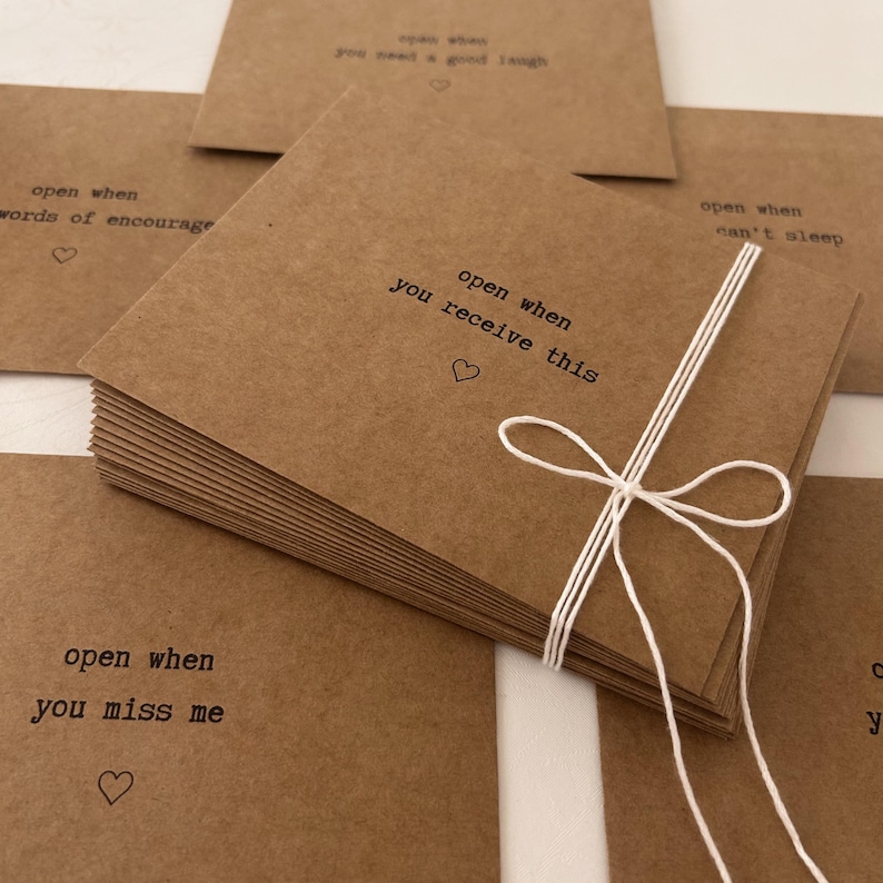 May include: A stack of brown kraft paper envelopes with white string tied around them. Each envelope has a different message printed on the front, such as "open when you receive this", "open when you miss me", and "open when you need a good laugh".
