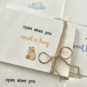 May include: A white card with a brown twine bow. The card has a watercolor illustration of two bears hugging and the text "open when you need a hug".