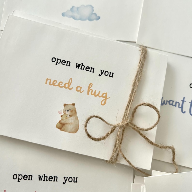 May include: A white card with a brown twine bow. The card has a watercolor illustration of two bears hugging and the text "open when you need a hug".