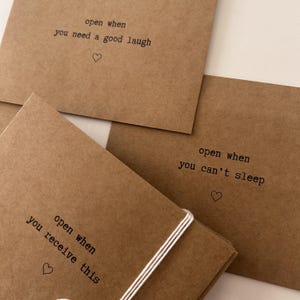 May include: Three brown envelopes with white string tied around them. Each envelope has a different message printed on the front: "open when you need a good laugh", "open when you receive this", and "open when you can't sleep".