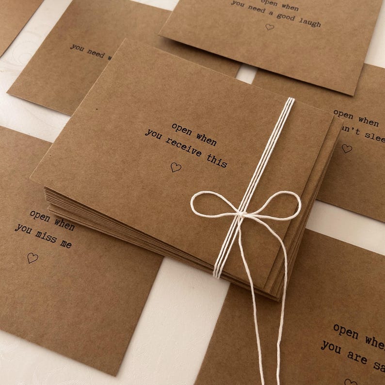 May include: A stack of brown envelopes tied with a white ribbon. Each envelope has a different message printed on the front, such as "open when you miss me" and "open when you need a good laugh".