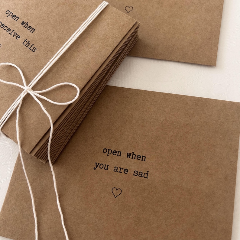May include: A stack of brown kraft paper envelopes with white string tied around them. Each envelope has the text "open when you are sad" printed on the front with a small heart symbol.