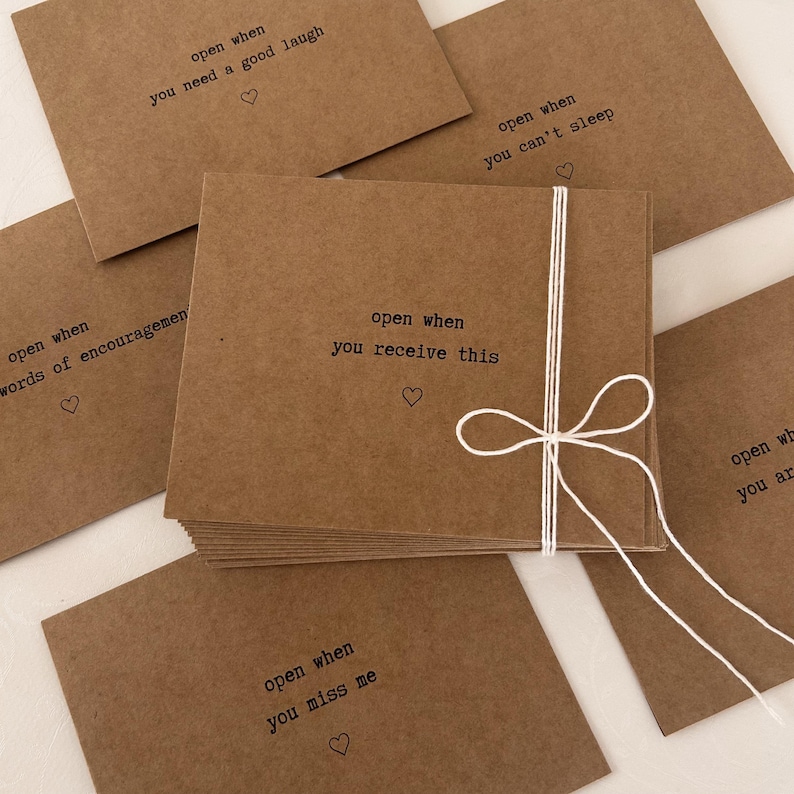 May include: A stack of brown envelopes with white string tied around them. Each envelope has a different message printed on the front, such as "open when you need a good laugh" and "open when you miss me".