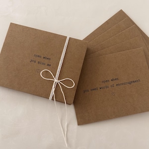 May include: A set of brown kraft paper cards tied together with white twine. Each card has a different message printed on it, such as "open when you miss me" and "open when you need words of encouragement".