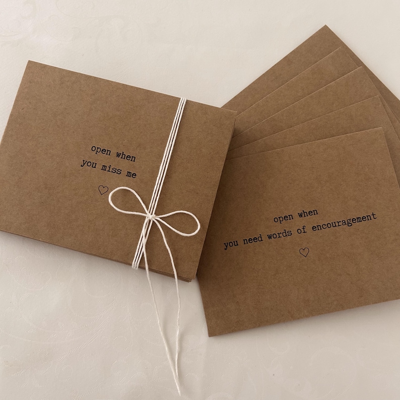 May include: A set of brown kraft paper cards tied together with white twine. Each card has a different message printed on it, such as "open when you miss me" and "open when you need words of encouragement".