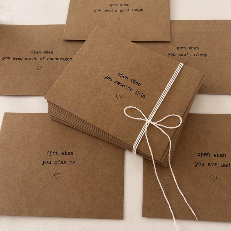 May include: A stack of brown envelopes with white string tied around them. Each envelope has a different message printed on the front, such as "open when you need a good laugh" and "open when you miss me".