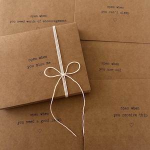 May include: A set of six brown kraft paper cards tied together with twine. Each card has a different message printed on it, such as "open when you need words of encouragement" and "open when you miss me".