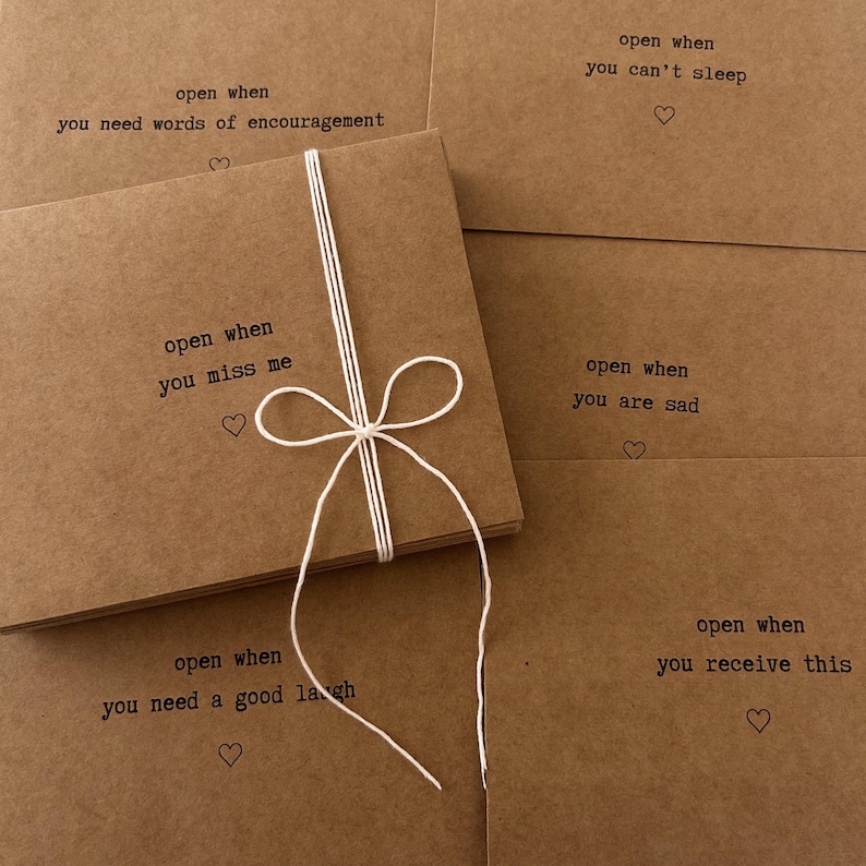 May include: A set of six brown kraft paper cards tied together with twine. Each card has a different message printed on it, such as "open when you need words of encouragement" and "open when you miss me".