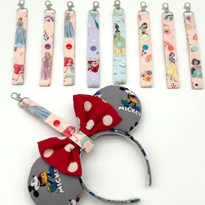 Puede incluir: Una colección de accesorios temáticos de personajes. Incluye una diadema gris con orejas de Mickey Mouse con un lazo rojo de lunares y varios llaveros de muñeca con diseños de princesas. Los llaveros tienen herrajes plateados y cierres a presión.