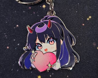 May include: A silver keychain with a charm featuring a cartoon character with purple hair, red horns, and a pink heart. The character is wearing a white shirt and has a light blue background.