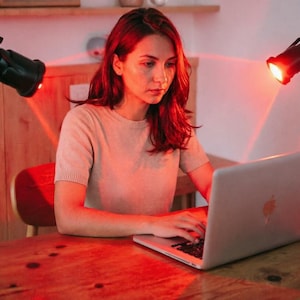 Könnte beinhalten: Eine Frau sitzt an einem Holztisch und benutzt einen silbernen Laptop. Die Szene wird von rotem Licht von zwei schwarzen Lampen beleuchtet. Sie trägt ein helles kurzärmeliges Oberteil. Der Tisch hat eine natürliche Holzmaserung.