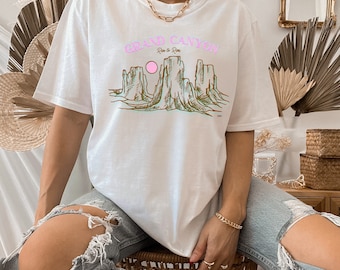 May include: White t-shirt with a graphic print of the Grand Canyon, featuring a pink sun and brown and green rock formations. The text "Grand Canyon" is printed in pink and blue letters with the words "Rim to Rim" printed in smaller letters below.