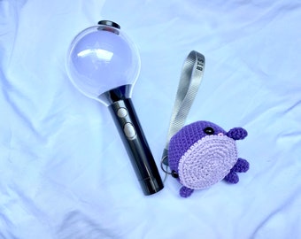 May include: A purple crocheted plush toy with black eyes and a clear plastic light-up stick with a black handle and a strap that says "BTS".