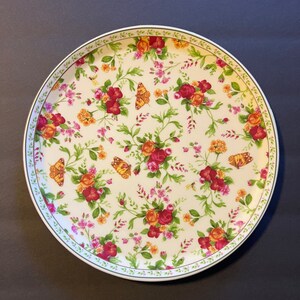 May include: A white ceramic plate with a floral pattern featuring red roses, green leaves, and orange butterflies. The plate has a green and white border.