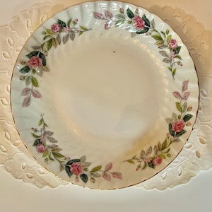 May include: A white porcelain plate with a gold rim and a floral design. The plate features a repeating pattern of pink roses, green leaves, and pink foliage around the rim. The plate has a scalloped edge.