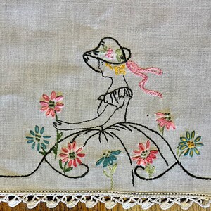 May include: White linen tablecloth with a crocheted edge. The tablecloth features an embroidered design of a girl in a hat and dress surrounded by flowers.