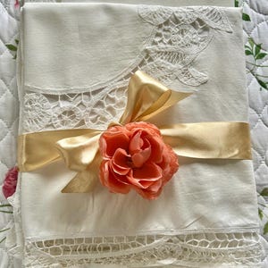 May include: Cream-colored linens with delicate lace trim are tied with a gold satin ribbon and adorned with a peach-colored rose. The linens are folded and appear to be new.
