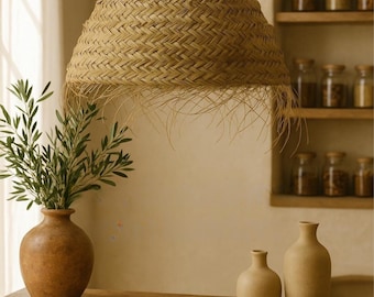 Op de afbeelding: Een geweven hanglamp van natuurlijke vezels hangt boven een houten tafel met drie keramische vazen en een kom. De lamp heeft een franje rand en is in een natuurlijke, lichtbruine kleur.