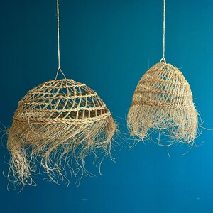 May include: Two woven pendant lights with a natural, beige color and a fringed bottom. The lampshades are suspended from the ceiling by a thin, off-white rope against a teal blue background. The lampshades have a bell-like shape.