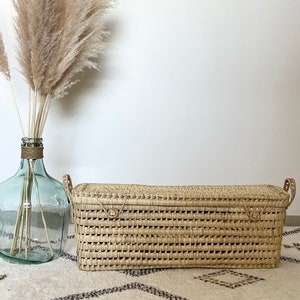 Puede incluir: Una cesta de almacenamiento rectangular tejida de forma natural con dos asas. La cesta está hecha de mimbre marrón claro y tiene un patrón tejido.